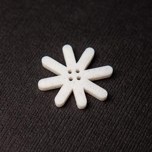 Puede incluir: Un botón blanco en forma de estrella con ocho brazos redondeados y cuatro pequeños agujeros en el centro. El botón tiene un aspecto moteado y brillante, sobre un fondo oscuro y texturizado.