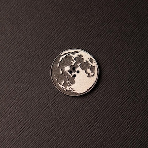 May include: A round, wooden button featuring a white moon design. The moon has a dark grey surface with craters and a white outline.