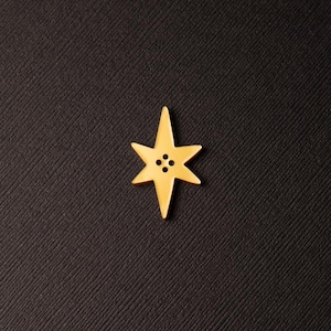 May include: A gold-colored star-shaped button with six points and four small holes in the center. The button is set against a dark, textured background, creating a contrast that highlights the button's shape and color.