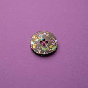 May include: A black button with a glittery iridescent finish. The button has four holes and is decorated with colorful confetti-like shapes.