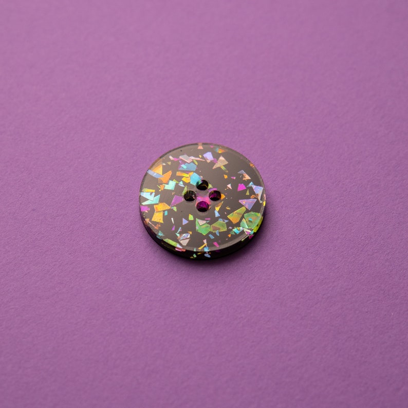 May include: A black button with a glittery iridescent finish. The button has four holes and is decorated with colorful confetti-like shapes.