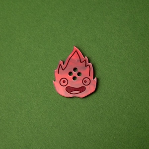 May include: Red plastic pin with a cartoon fire creature design. The creature has a smiling face with two eyes and a mouth. There are four small circles on the creature's body.