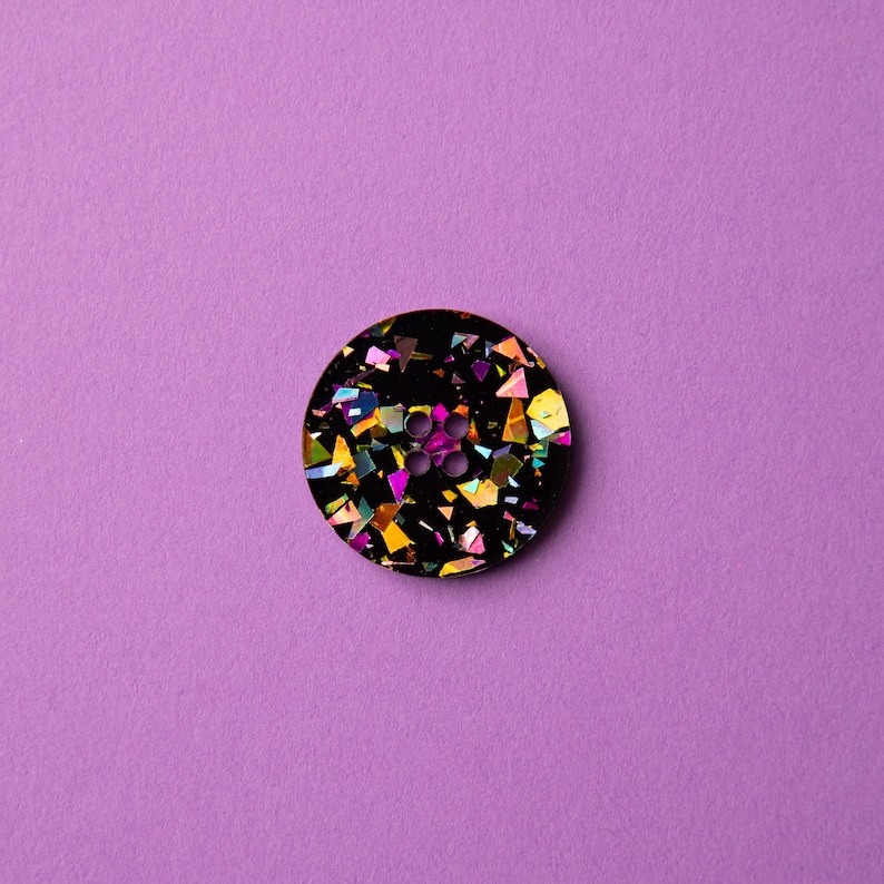 May include: A black button with a four-hole design and iridescent glitter embedded in the resin.