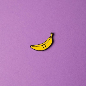 May include: A yellow banana pin with black dots on a purple background.