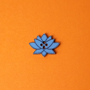 May include: A light blue lotus flower-shaped button with three holes, set against an orange background. The button has a detailed outline and is likely made of wood or a similar material. This decorative button could be used for sewing or crafts.