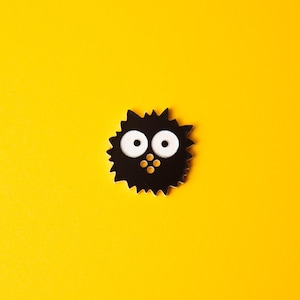 May include: A black, spiky character with large white eyes and three small yellow dots below. The figure is set against a bright yellow background, creating a striking contrast.