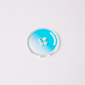 May include: A round, clear button with a gradient of white to turquoise blue. Four small, silver-colored embellishments are in the center. The button has a glossy finish and is set against a white background.