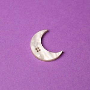 May include: A white crescent moon-shaped button with four small holes in the centre, on a purple background.