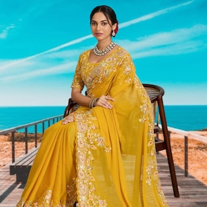 Puede incluir: Una mujer con un sari amarillo mostaza con intrincados bordados y adornos. El sari presenta un borde floral detallado y se combina con una blusa a juego. Está sentada en una silla de madera con un fondo de mar y cielo.