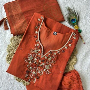 Könnte beinhalten: Ein orangefarbenes, besticktes Kurta-Set mit passendem Dupatta. Das Kurta hat aufwendige Blumenstickereien um den Ausschnitt und den Oberkörper. Das Dupatta hat einen goldenen Rand und Quasten. Eine Pfauenfeder ist in der Nähe platziert.