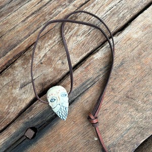 May include: A handcrafted pendant necklace with a carved, off-white face pendant. The pendant has blue eyes and a patterned beard, suspended from a brown leather cord. The necklace is knotted for adjustable length, and lies on a wooden surface.
