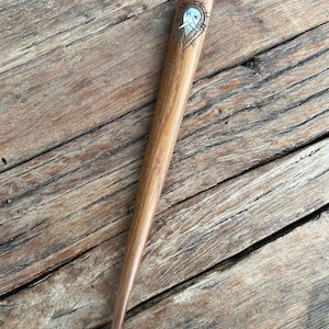 May include: A wooden hair stick with a tapered design. The top of the stick features an engraved design of a face with a beard and a light blue background. The hair accessory is made of a light brown wood.