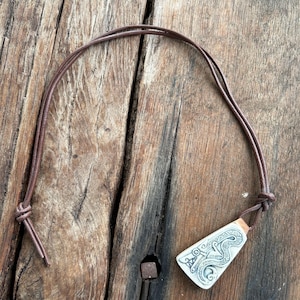 May include: A pendant necklace with a rectangular, off-white pendant featuring an engraved design. It hangs from a brown leather cord. The necklace is displayed on a weathered wooden surface.
