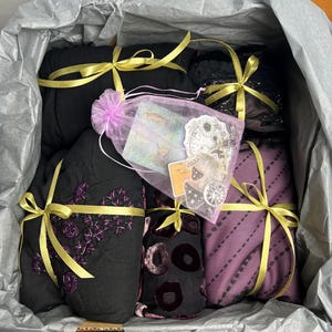 May include: A gift box containing wrapped items in black and purple fabrics, tied with gold ribbons. A small, sheer, pink bag with cards and stickers is also included. The box is lined with grey tissue paper. The items are in various patterns.