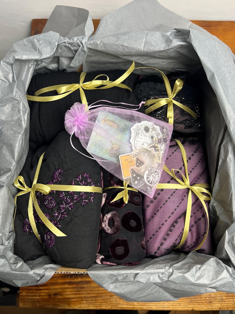 May include: A gift box containing wrapped items in black and purple fabrics, tied with gold ribbons. A small, sheer, pink bag with cards and stickers is also included. The box is lined with grey tissue paper. The items are in various patterns.