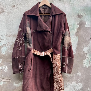 Op de afbeelding: Een bruin corduroy jack met een riem en geborduurde details. Het jack heeft een patchwork ontwerp met verschillende stoffen en texturen.
