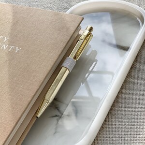 Könnte beinhalten: Ein ovales Tablett aus weißem Marmor mit einem goldenen Stift und einem braunen Notizbuch darauf. Das Tablett hat einen weißen Rand und steht auf einer beigefarbenen Stoffoberfläche.