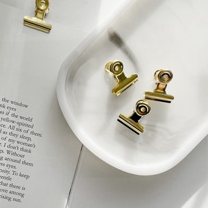 Pode incluir: Três clipes de pasta dourados sobre uma superfície de mármore branco. Os clipes são um material de escritório comum usado para prender papéis.