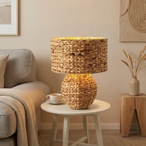 Puede incluir: Una lámpara de mesa con pantalla cilíndrica y base redondeada, ambas fabricadas con fibras de pasto marino tejidas. La lámpara emite un brillo cálido y suave desde su interior, resaltando la intrincada textura del material trenzado.
