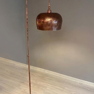 Op de afbeelding: Een vloerlamp met een hoge, dunne, koperkleurige metalen standaard en een bijpassende ronde voet. De lamp heeft een grote, koepelvormige kap met een getextureerde, gehamerde koperen afwerking. Een zwart snoer ligt opgerold op de voet van de lamp.