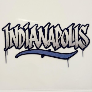 May include: A sticker with the word "INDIANAPOLIS" in a graffiti-style font. The text is white with a gray gradient and a dark blue outline. A blue curved line is below the text, with blue drips.