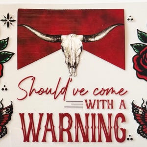 Pode incluir: Uma coleção de desenhos em estilo tatuagem com um crânio de longhorn, borboletas em chamas, rosas e folhas. O texto "Should've come with a warning" é exibido em vermelho. O design está em um fundo branco.