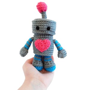 May include: A gray crocheted robot with blue and white striped arms and legs. The robot has a pink heart on its chest and a pink pom-pom on its head.
