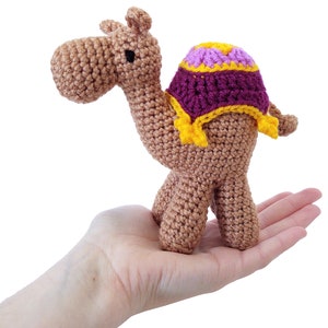 Peut inclure: Un jouet en crochet en forme de chameau avec une selle violette et jaune. Le chameau est brun et a une tête amicale.