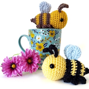 May include: Two crocheted bumblebees, one yellow and black, the other yellow and brown, sitting on a blue mug with a floral pattern. The mug is on a white surface.