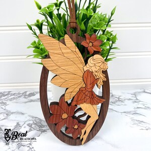 May include: Wooden fairy ornament with a brown oval frame, featuring a fairy with light brown wings and a red-brown dress. The ornament includes two brown flowers and a leather cord for hanging. The text "Real Creations" is at the bottom.
