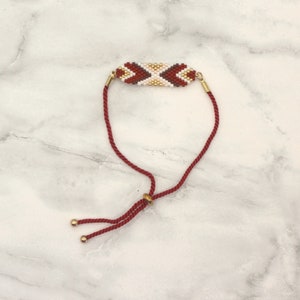 May include: A red and white beaded bracelet with a geometric pattern. The bracelet is adjustable with a sliding knot and gold-toned beads.