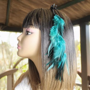 May include: A teal feather hair clip attached to a black hair accessory. The feathers are a vibrant teal color and cascade down the side of the head. The hair clip is attached to a mannequin head with long, dark hair.