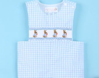 Peut inclure: Salopette bébé en vichy bleu et blanc avec un motif brodé de Lapin de Pâques. La salopette est sans manches et se ferme par un bouton. Un œuf de Pâques en crochet rose et vert est en arrière-plan.