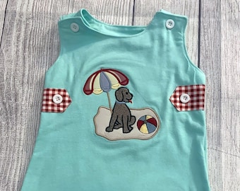 Peut inclure: Une barboteuse turquoise sans manches avec une bordure en vichy rouge et blanc. La barboteuse présente un motif brodé d'un chien assis sur une plage avec un parasol et un ballon de plage.