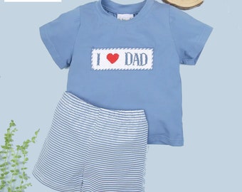 Peut inclure: Un t-shirt bleu à manches courtes avec un patch brodé blanc et rouge qui dit "J'aime Papa". Le t-shirt est associé à un short rayé bleu et blanc.