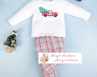 May include: White long-sleeved shirt with a red truck carrying a green Christmas tree. Red and green plaid trousers with a white background. The text "High Cotton Magnolias" is on the trousers.