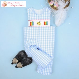 May include: Blue and white gingham overalls with a white embroidered bib featuring a rainbow of crayons, paintbrushes, and a paint palette. The overalls are on a light blue background.