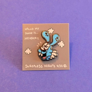 Könnte beinhalten: Eine blaue Emaille-Anstecknadel mit einer Comicfigur mit einem Auge, einem breiten Lächeln und dem Text "HELLO MY NAME IS... HERBERT." und "SLEEPELESS NIGHTZ 437 ©".