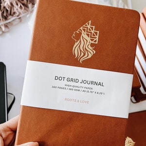 Peut inclure: Un carnet à points de grille relié en cuir marron avec un motif en relief doré représentant le visage d'une femme avec une couronne. Le carnet porte l'inscription "DOT GRID JOURNAL", "HIGH QUALITY PAPER", "160 PAGES/160 GSM/A5 (5.75" X 8.25")", et "ROOTS & LOVE" imprimée sur la couverture.