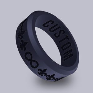 May include: A black silicone ring with the word "CUSTOM" engraved on the side and a repeating pattern of fleur-de-lis and infinity symbols.