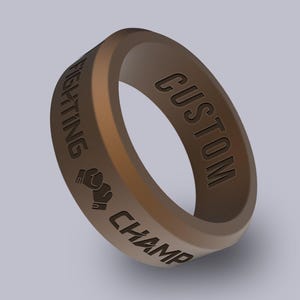 May include: A brown silicone ring with the words "CUSTOM FIGHTING CHAMP" engraved around the band. A boxing glove graphic is also present. The ring is a solid color with a smooth finish.