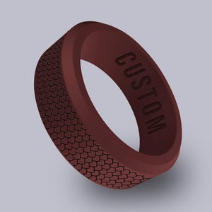May include: A maroon silicone ring with a textured, scale-like pattern on the outer band. The word "CUSTOM" is engraved on the ring's surface. The ring is a simple band design.