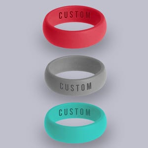 May include: Three silicone rings in red, gray, and teal, each with the word "CUSTOM" embossed on the band. The rings are displayed against a light purple background, showcasing their colors and design.