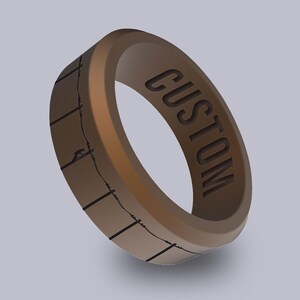 May include: A brown ring with the word "CUSTOM" engraved on the inside. The ring has a textured design with black lines and a grid pattern on the outside.