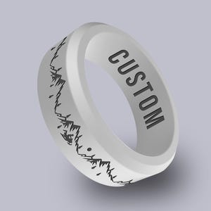 May include: A white silicone ring with a mountain range design in black. The word "CUSTOM" is printed in black on the ring. The ring is a simple band, suitable for everyday wear.