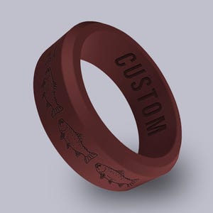 May include: A dark maroon silicone ring featuring a repeating pattern of black line-art fish engravings around the outer band. The word CUSTOM is debossed in black capital letters on the side of the ring, which has a smooth, matte finish.