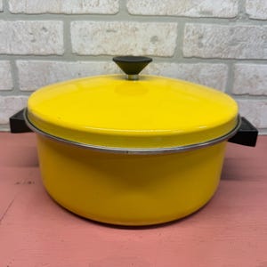 May include: A bright yellow enamel pot with a lid and black handles. The pot has a silver rim and a black knob on the lid. The pot is sitting on a pink surface in front of a white brick wall. Kitchenware.