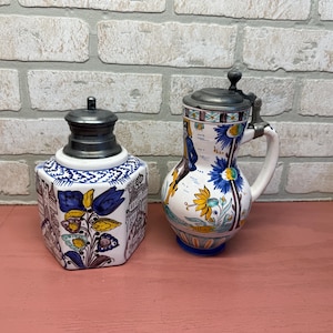 May include: Two antique ceramic pieces: a lidded inkwell and a beer stein. The inkwell is hexagonal with a floral design and a metal lid. The stein has a handle, a hinged metal lid, and is decorated with flowers and figures.