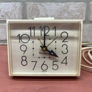 May include: A white vintage Westclox alarm clock with black numbers and hands. The clock has a white cord and is made in the USA.