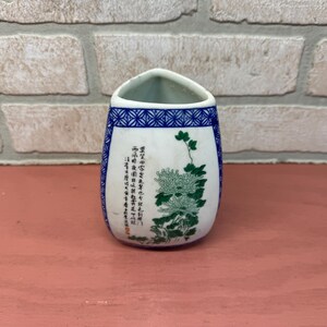 May include: A small, white ceramic vase with a blue and green floral design. The vase has a triangular shape and is decorated with a floral pattern and Chinese characters.